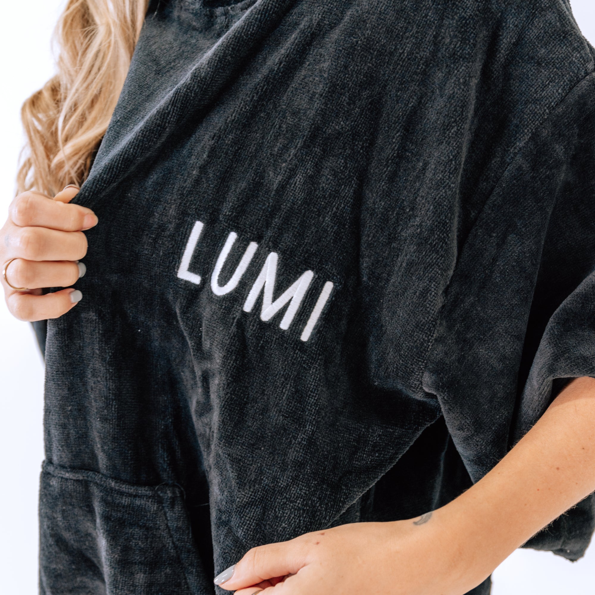 Close-up view of LUMI Towel Poncho showing plush black cotton fabric with white embroidered LUMI logo on chest, demonstrating texture and quality of material, with side pockets visible against dark background
