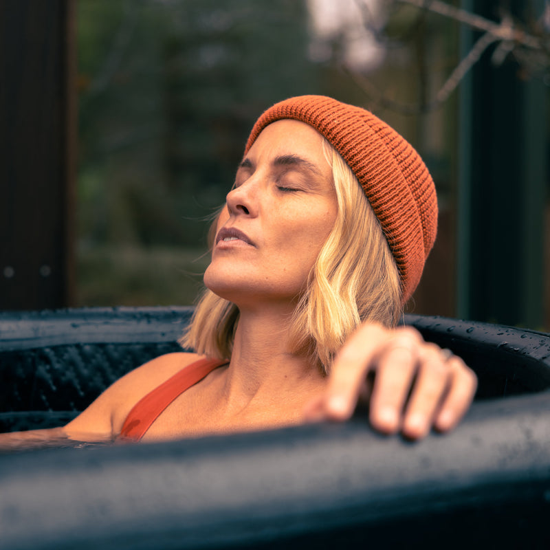 Photo of a woman wearing an orange hat and swimsuit, sat in the LUMI Pro Duo ice bath, eyes closed and focused on recovery.