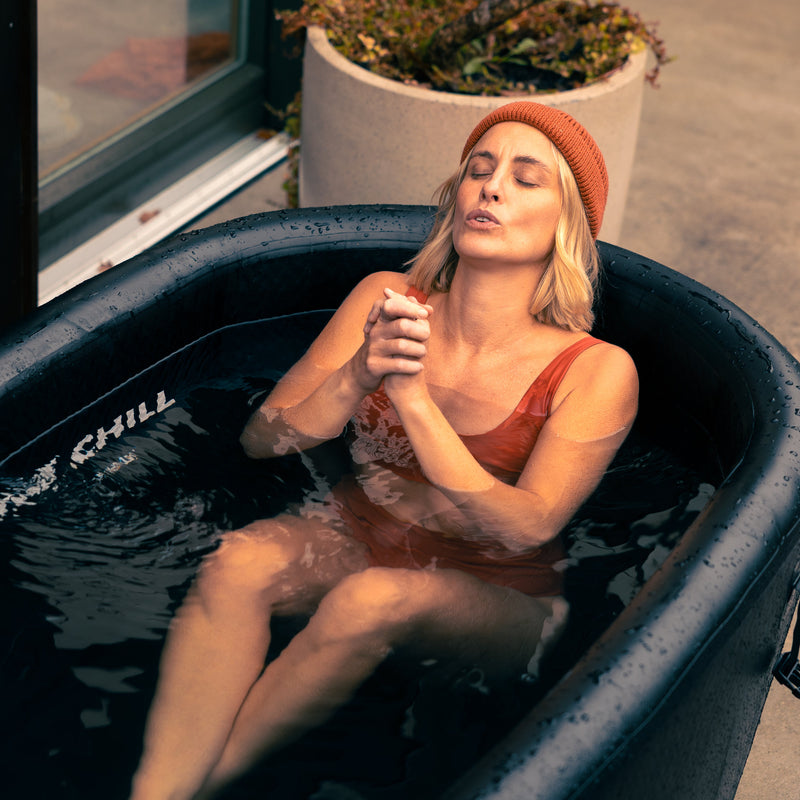 Photo of a woman wearing an orange hat and swimsuit, in the LUMI Pro Duo ice bath, eyes closed with hands clasped, focused on cold water recovery.