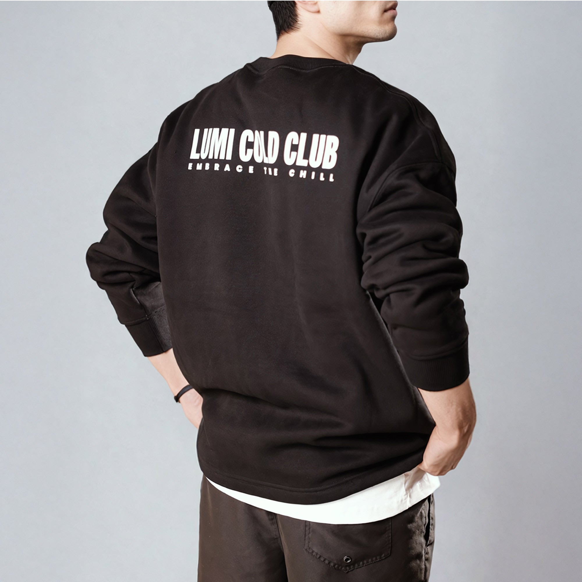 Back view of Unisex LCC Oversized Sweatshirt Black showing white LUMI Cold Club text graphic, worn over white undershirt, demonstrating oversized fit and premium cotton-polyester fabric quality in a comfortable, casual styling.