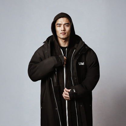 PRO DUO Changing Robe in black, featuring a modern winter jacket design with hood, windproof sleeves, and LUMI logo. Studio photo showing full-length waterproof robe with contrasting white zipper details against dark fabric, demonstrating the roomy yet stylish Nordic-inspired silhouette.