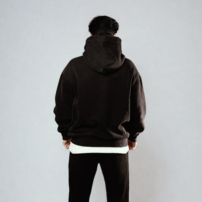 Back view of Unisex LUX Oversized Hoodie Black Wash shown on figure against white background, featuring relaxed fit, dropped shoulders, and layered white underlining at hem, demonstrating the hoodie's oversized silhouette and premium casual style.