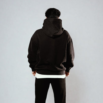 Back view of Unisex LUX Oversized Hoodie Black Wash shown on figure against white background, featuring relaxed fit, dropped shoulders, and layered white underlining at hem, demonstrating the hoodie's oversized silhouette and premium casual style.