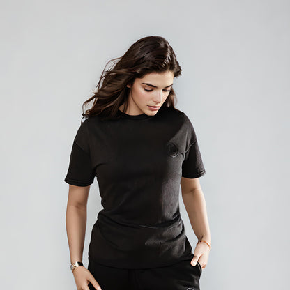 Model wearing Women's LUX T-Shirt Black Wash, a premium cotton heavyweight tee with classic fit and subtle rubber logo detail, styled casually with one hand in pocket against light grey background, showcasing the shirt's relaxed silhouette and washed black color.