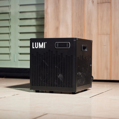 LUMI Cube Chiller compact black cooling unit for ice baths positioned on light tile flooring against wooden paneling and mint green shutters, showcasing its sleek design with ventilated sides and digital display screen