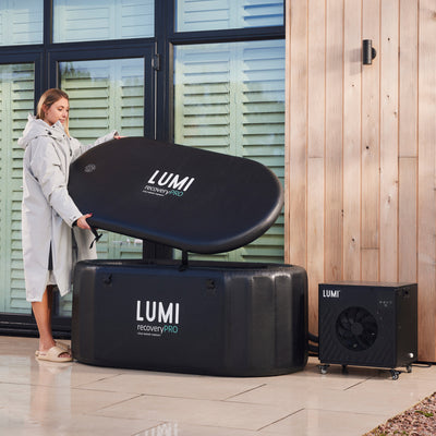 The Cube PRO Chiller shown alongside a LUMI recovery pod system, featuring a sleek black cooling unit and padded ice bath container on an outdoor patio with wooden wall paneling and shuttered windows. Professional recovery equipment setup demonstrates complete cold therapy system installation.