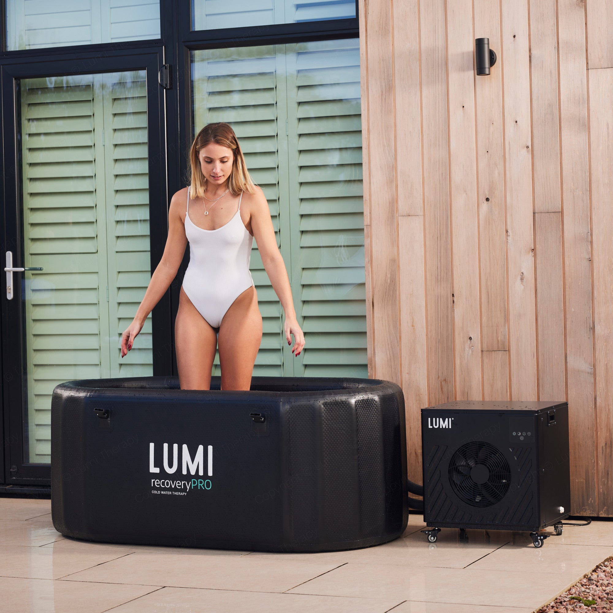 LUMI Recovery Pro ice bath setup with Cube PRO Chiller system featuring a black portable tub and compact chiller unit positioned on light wooden decking against mint green shutters and natural wood paneling.