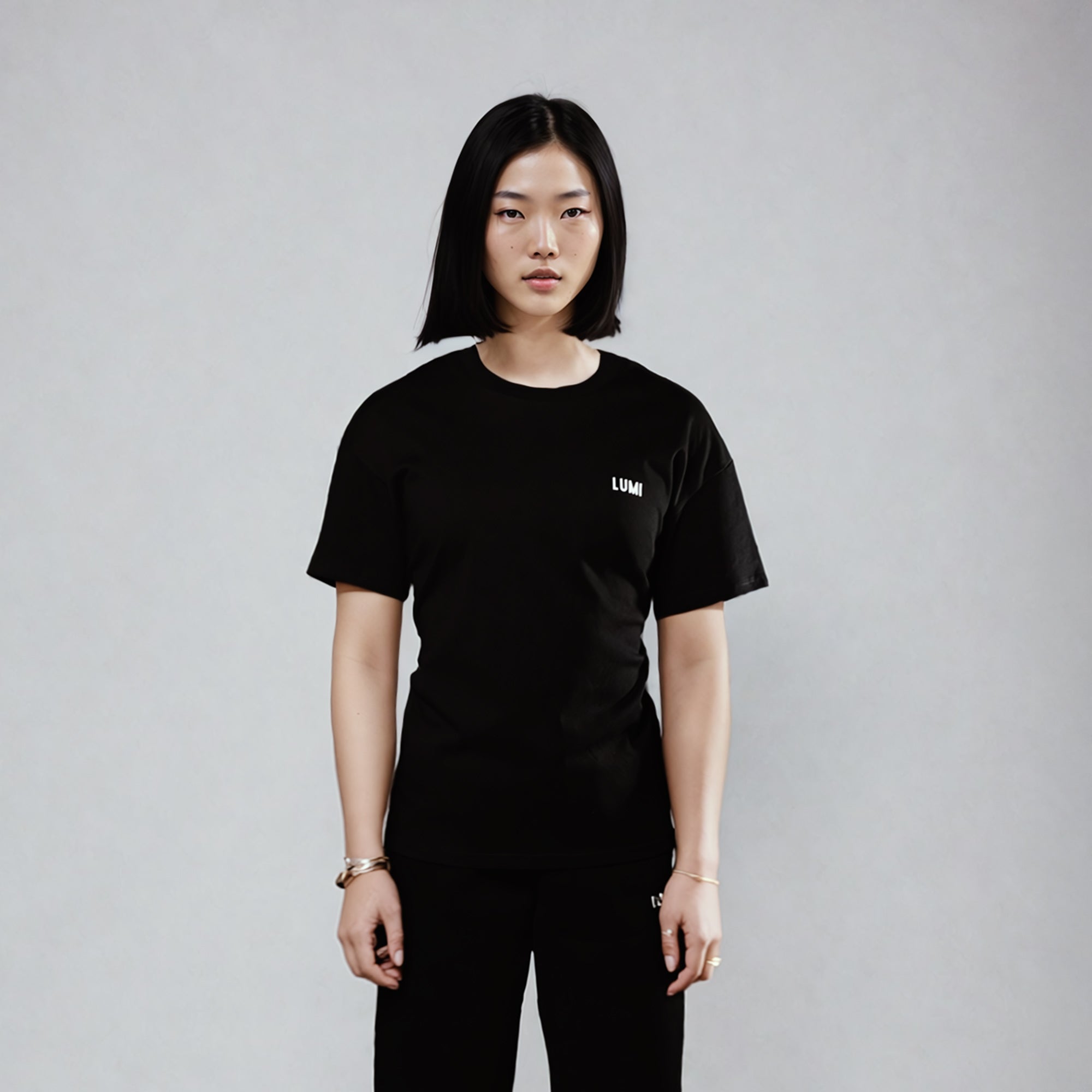 Women's LCC T-Shirt Black shown in a minimalist studio setting against a gray backdrop, featuring a classic crew neck design with short sleeves and small white LUMI logo on chest. Heavyweight cotton t-shirt styled with black pants for a monochromatic look.