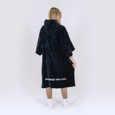 LUMI Towel Poncho shown from back view in deep teal plush fabric, featuring hooded design, mid-calf length, and 'Embrace the Chill' text detail at hem, styled with white socks and displayed on light background