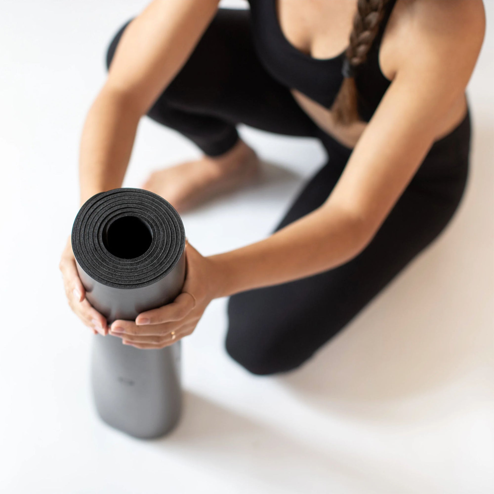 A person in black athletic wear rolling up an Eco Wolf Yoga Mat - Nude Edition, shown from above against a white background. The mat appears in a dark charcoal color, demonstrating its portability and compact rolling design.