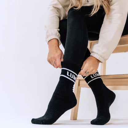 Pro Bamboo Socks - With Stripes (4 Pack) shown on feet against white background, featuring black athletic socks with white LUMI branding and stripe detail. Model seated on wooden chair wearing cream-colored top and black pants, demonstrating sock fit and style.