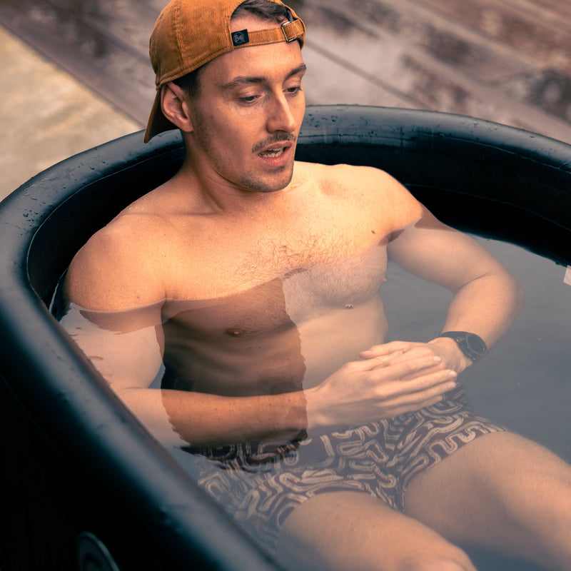 Photo of a man wearing a baseball cap and swim shorts, sitting in the LUMI Pro Duo ice bath, focused on cold water recovery.