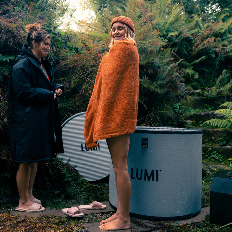 Two women stood beside the LUMI Pod Pro ice bath after a cold water therepy session, one wrapped in an orange towel and the other wearing the LUMI RobePRO DUO Changing Robe.