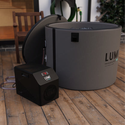 LUMI Mini Chiller Black Edition shown with its compact cooling unit next to a large circular ice bath tub on wooden deck flooring. The sleek black and grey design features modern styling and digital display for temperature control.