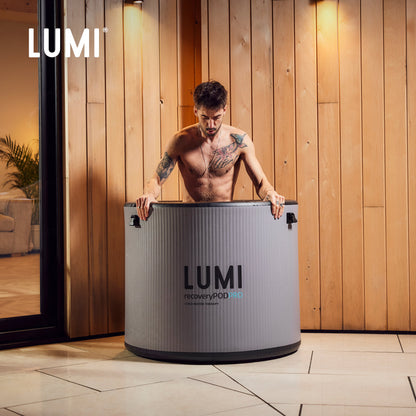 Pod PRO Ice Barrel shown in modern wellness setting with warm wood paneling background. The stone grey cylindrical barrel features sleek design and LUMI branding, demonstrating its use for cold therapy immersion against elegant interior lighting.